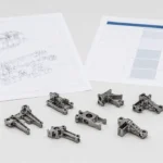 Escena de escritorio de ingeniería con notas de comparación de materiales MIM, un plano de pieza y piezas pequeñas de moldeo por inyección de metal preparadas para la revisión del proveedor.